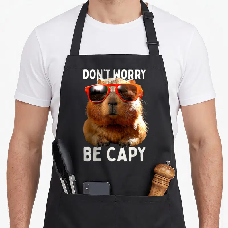 Tablier de cuisine noir avec un cochon d'eau portant des lunettes de soleil orange et le texte Don't Worry Be Capy.