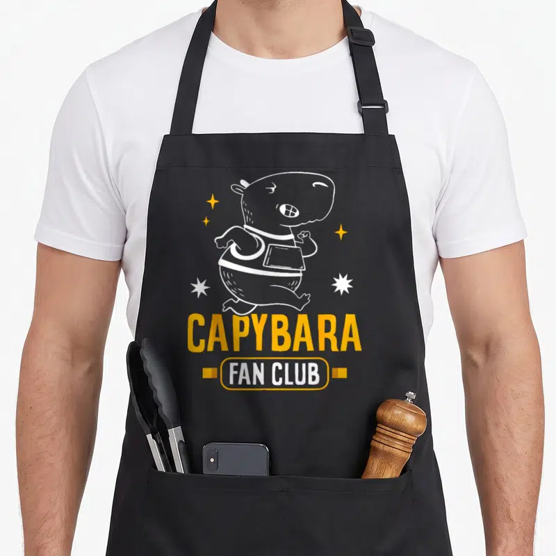 Tablier de cuisine noir avec illustration blanche d'un capybara dansant et le texte CAPYBARA FAN CLUB en orange.