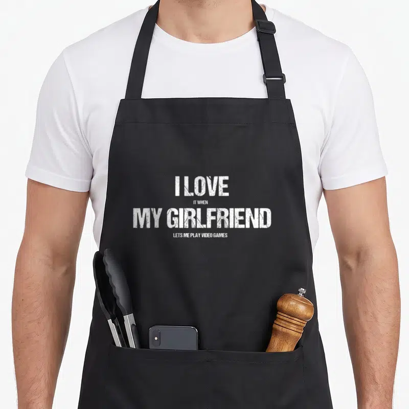 Tablier de cuisine noir avec le texte blanc I Love My Girlfriend Lets Me Play Video Games imprimé au centre.