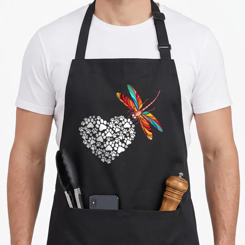 Tablier de cuisine noir avec une libellule multicolore et un cœur composé de pattes de chien blanches.