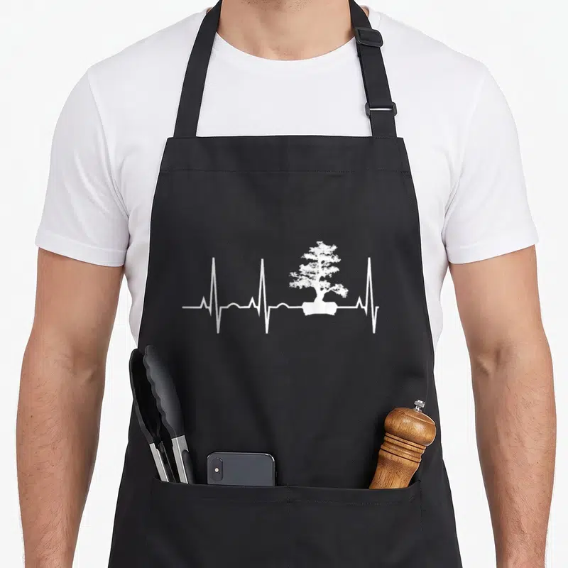 Tablier de cuisine noir avec un motif blanc représentant un bonsai au centre d'une ligne de rythme cardiaque.