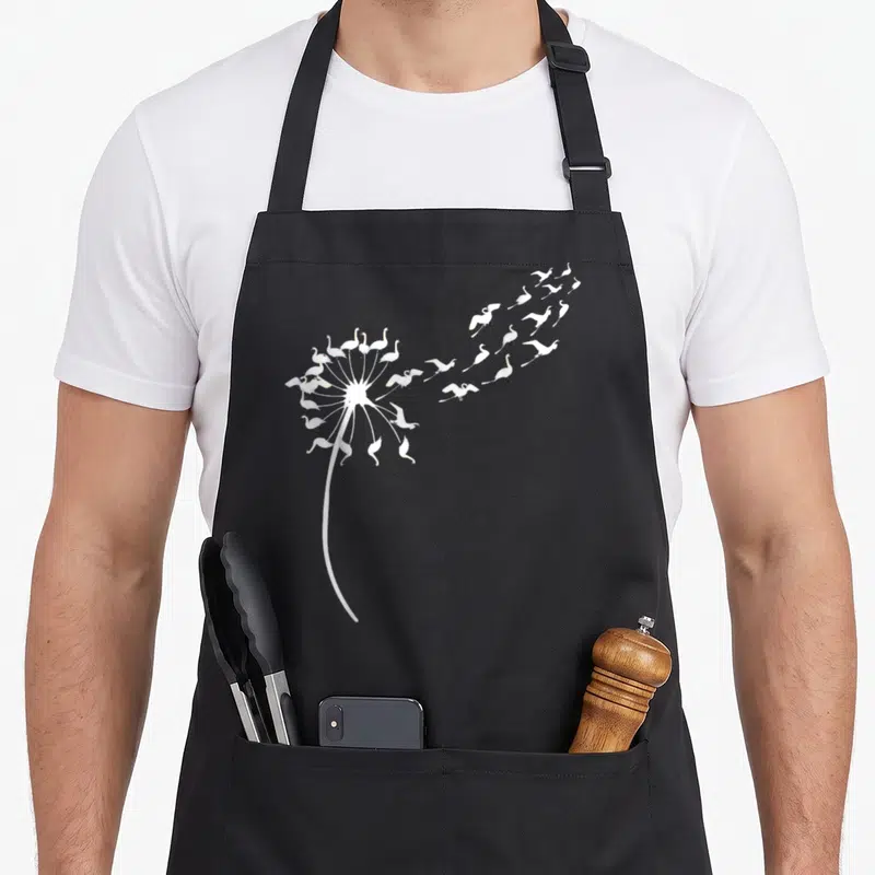 Tablier de cuisine noir avec une illustration d'un pissenlit dont les graines sont des cygnes blancs en vol.