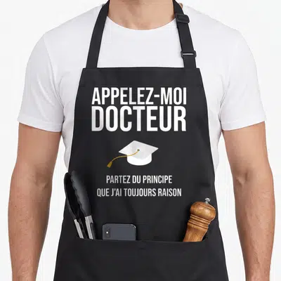 Tablier de cuisine noir avec le texte Appelez-moi docteur au-dessus d'une toque blanche stylisée et d'une phrase humoristique en blanc.