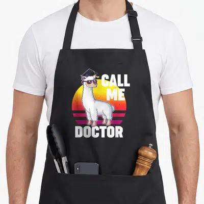 Tablier de cuisine noir avec illustration d'un lama en toque et lunettes de soleil devant un soleil couchant coloré.