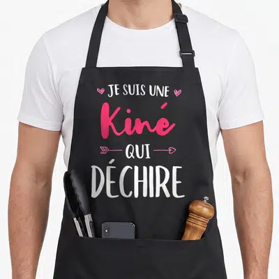 Tablier de cuisine noir avec l'inscription Je Suis Une Kiné Qui Déchire en rose et blanc, orné de cœurs et d'une flèche.