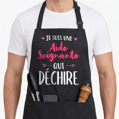Tablier de cuisine noir avec inscription rose et blanche Je suis une aide soignante qui déchire ornée de cœurs et d'une flèche.