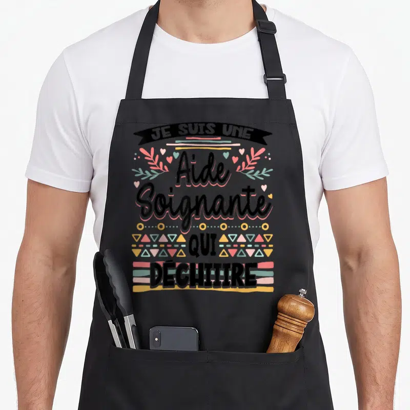 Tablier de cuisine noir avec inscription Je suis une aide soignante qui déchire entourée de motifs géométriques et de cœurs pastel.