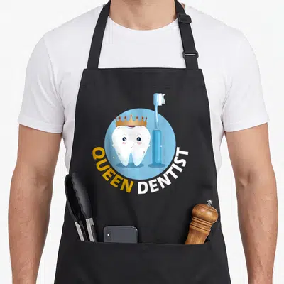 Tablier de cuisine noir avec une dent souriante coiffée d'une couronne et une brosse à dents bleue sur fond rond.