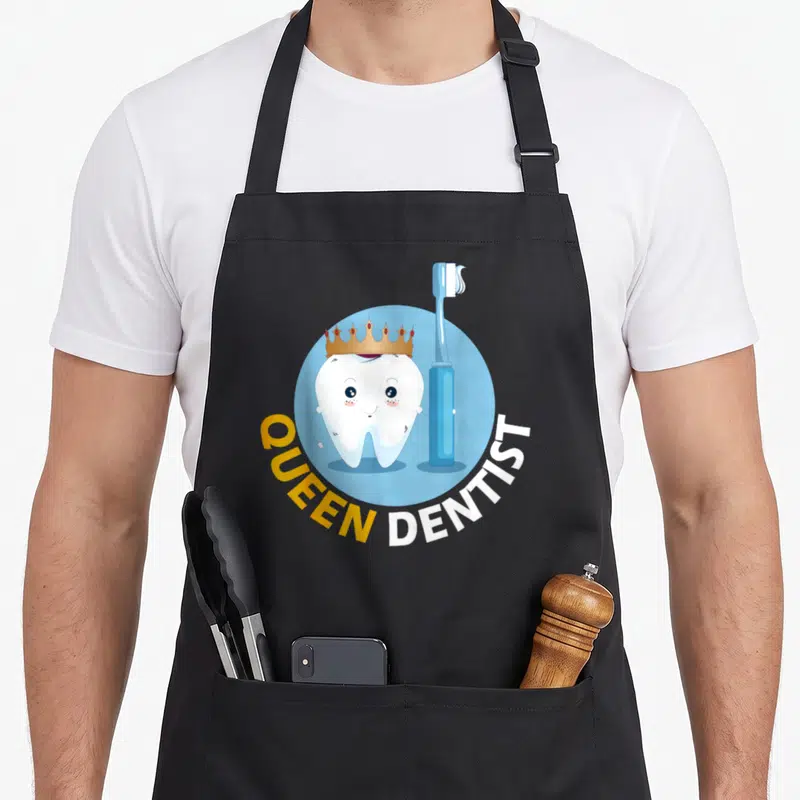 Tablier de cuisine noir avec une dent souriante coiffée d'une couronne et une brosse à dents bleue sur fond rond.