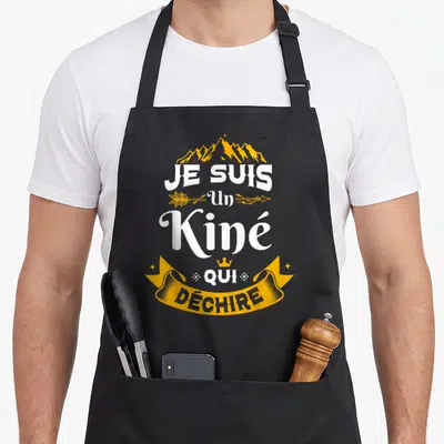 Tablier de cuisine noir avec l'inscription Je suis un Kiné qui déchire sur fond de montagnes dorées et une bannière stylisée.