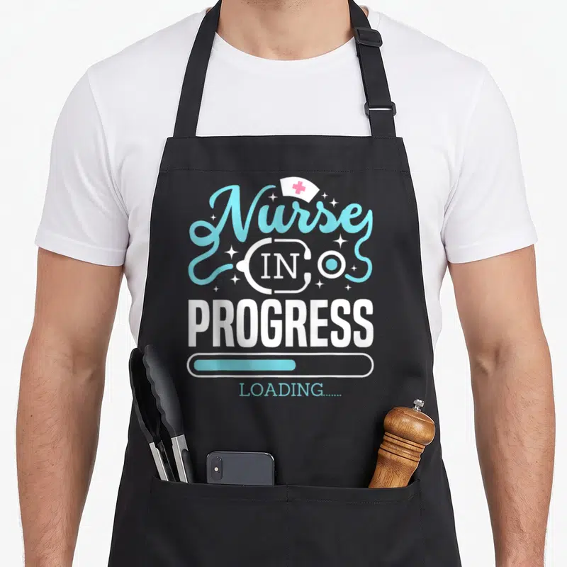 Tablier de cuisine noir avec l'inscription Nurse in Progress Loading et une barre de chargement bleue.