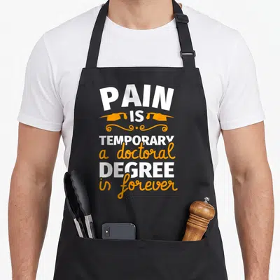 Tablier de cuisine noir avec l'inscription blanche et orange « PAIN IS TEMPORARY a doctoral DEGREE is forever » surmontée de deux bonnets de graduation.