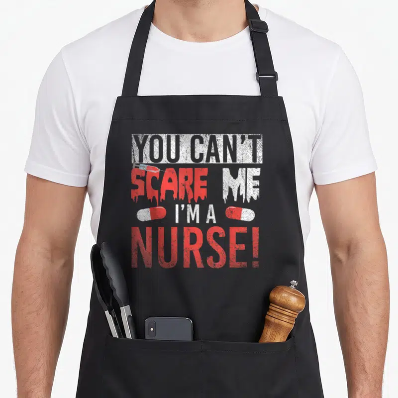 Tablier de cuisine noir avec texte You Can't Scare Me I'm A Nurse, seringue et deux pilules rouges et blanches.