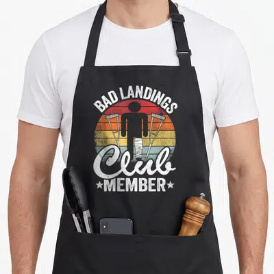 Tablier de cuisine noir avec illustration rétro d'une personne aux béquilles et jambe en plâtre sur fond arc-en-ciel délavé.