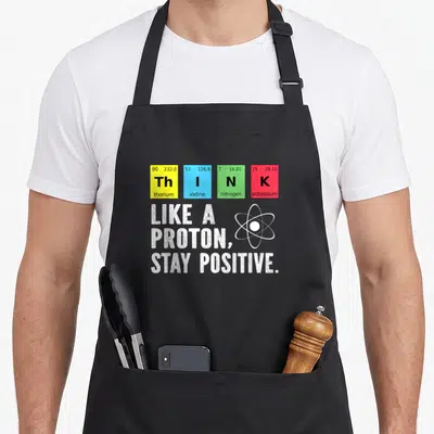 Tablier de cuisine noir avec les symboles chimiques ThINk et la phrase Like a Proton Stay Positive accompagnée d'un schéma atomique.