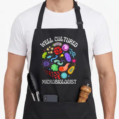 Tablier de cuisine noir avec illustrations colorées de microbes et le texte Well Cultured Microbiologist en blanc.