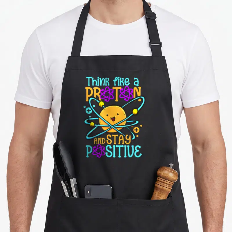 Tablier de cuisine noir avec un proton souriant entouré d'orbites atomiques et du texte Think like a Proton And Stay Positive.