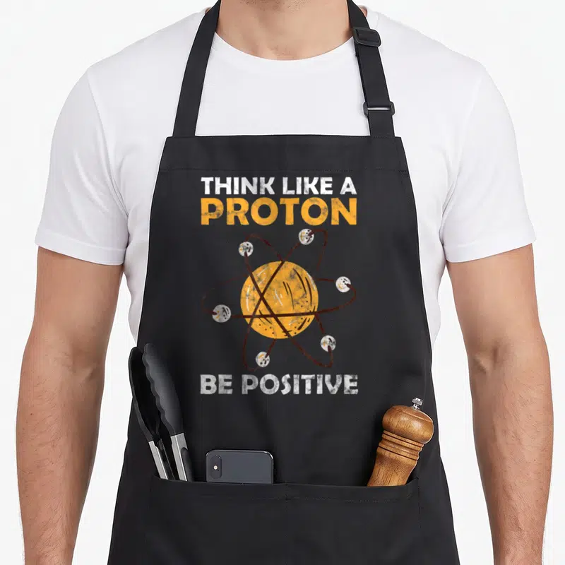 Tablier de cuisine noir avec illustration d'atome orange et texte blanc Think Like A Proton Be Positive