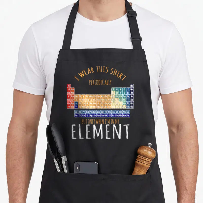 Tablier de cuisine noir avec un tableau périodique coloré et des phrases en anglais humoristiques sur fond gris uni.