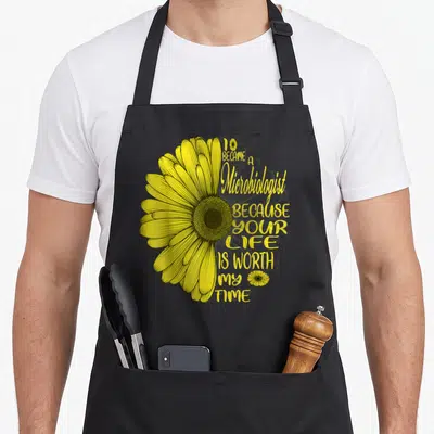 Tablier de cuisine noir avec une illustration de marguerite jaune et le texte « I became a Microbiologist because your life is worth my time ».