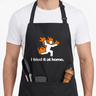 Tablier de cuisine noir avec un personnage blanc en feu et le texte I tried it at home imprimé en bas.