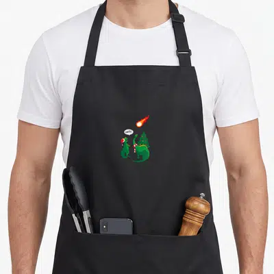 Tablier de cuisine noir avec deux dinosaures verts et un météore rouge en illustration centrale.