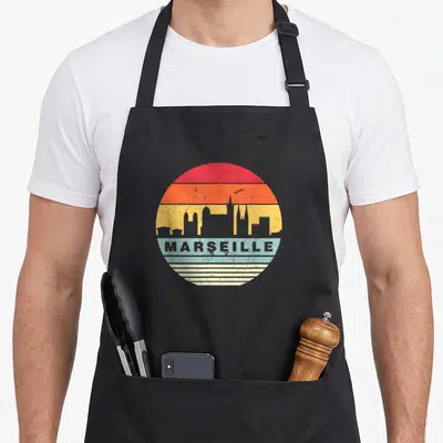 Tablier de cuisine noir avec un motif circulaire coloré représentant la silhouette de Marseille sur fond de coucher de soleil rétro.