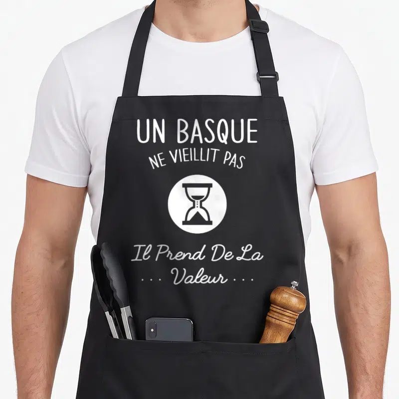 Tablier de cuisine noir avec texte blanc Un Basque ne vieillit pas et illustration d'une horloge à sable.