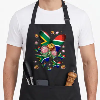 Tablier de cuisine noir avec papillons au drapeau sud-africain, fleurs de protea et mets typiques imprimés sur le devant.