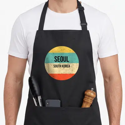 Tablier de cuisine noir avec un motif circulaire coloré affichant les mots Séoul South Korea sur des bandes horizontales.