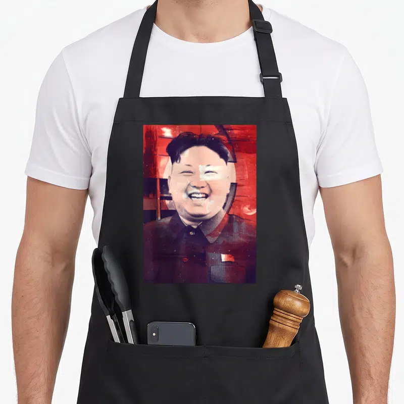 Tablier de cuisine noir avec portrait stylisé rouge et blanc de Kim Jong Un souriant sur fond abstrait.