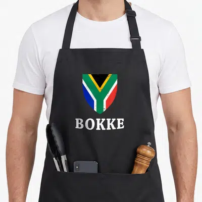 Tablier de cuisine noir avec un bouclier aux couleurs du drapeau sud-africain et le texte BOKKE en blanc.