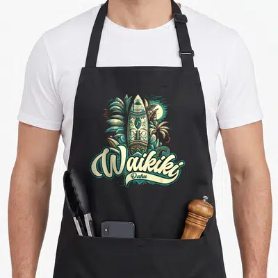 Tablier de cuisine noir avec une planche à vagues ornée de motifs tribaux, de palmes et l'inscription Waikiki Oahu.