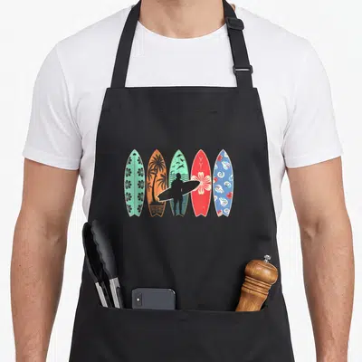Tablier de cuisine noir avec cinq planches de surf colorées et une silhouette de surfeur tenant une planche noire.