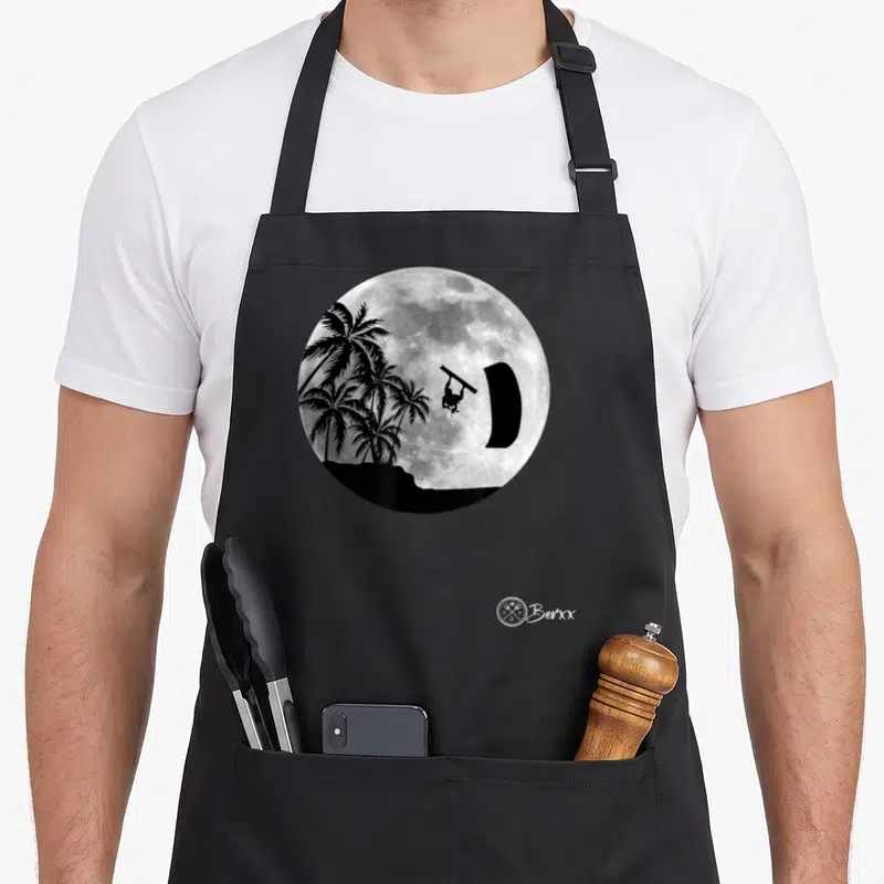 Tablier de cuisine noir avec une lune pleine, des palmiers et un kitesurfeur en silhouette noire.