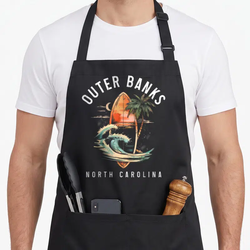 Tablier de cuisine noir avec planche de surf orange, palmier et vague turquoise sous le texte Outer Banks North Carolina.