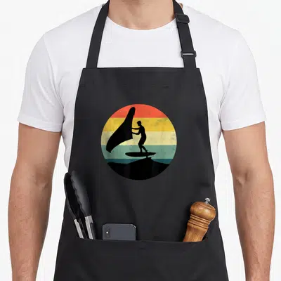 Tablier de cuisine noir avec silhouette d'un wingfoiler tenant une aile sur fond de bandes colorées rondes.