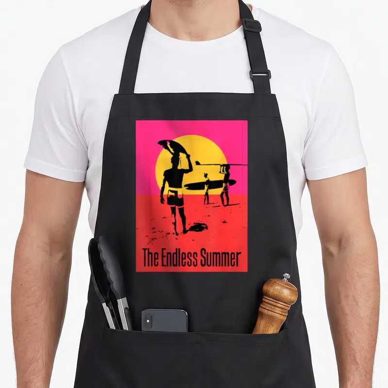 Tablier de cuisine noir avec affiche surf The Endless Summer, silhouettes et soleil orange sur fond rose.