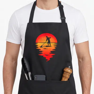 Tablier de cuisine noir avec silhouette de paddle sur fond de soleil couchant orange et reflet dans l'eau.