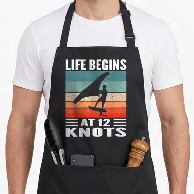 Tablier de cuisine noir avec silhouette wing foil et texte blanc Life Begins At 12 Knots sur bandes colorées vintage.