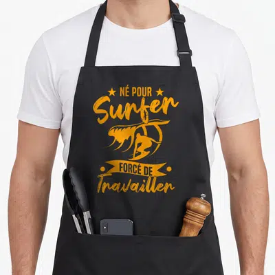 Tablier de cuisine noir avec inscription orange « Né pour Surfer Forcé de Travailler » et silhouette de surfeur sur une vague.