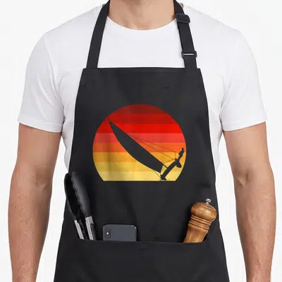 Tablier de cuisine noir avec illustration circulaire d'un catamaran au sunset rouge orange jaune
