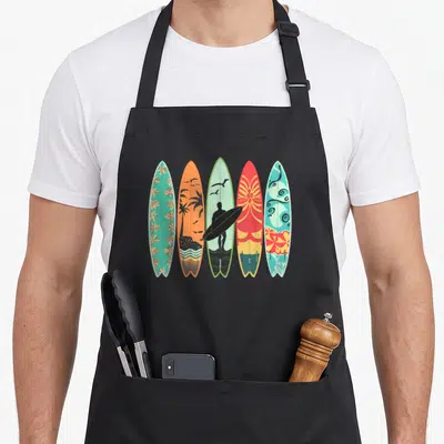 Tablier de cuisine noir avec cinq planches de surf aux motifs colorés et floraux.