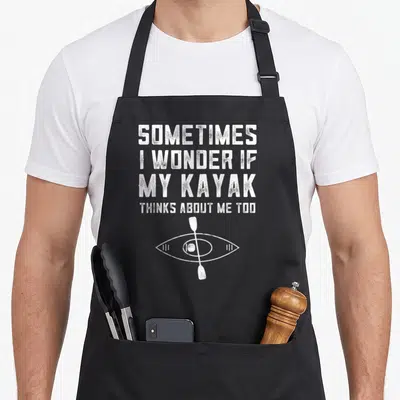 Tablier de cuisine noir avec le texte Sometimes I Wonder If My Kayak Thinks About Me Too et un dessin d'œil traversé par une paddle.