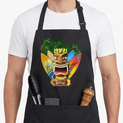 Tablier de cuisine noir avec un tiki hawaïen entouré de planches de surf et de fleurs tropicales colorées.