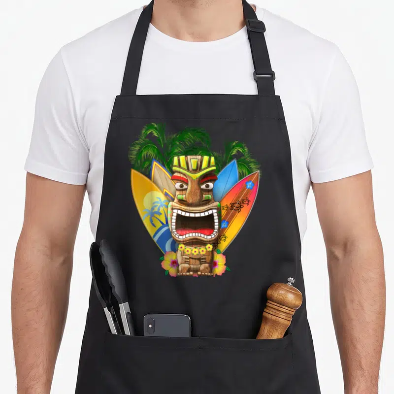 Tablier de cuisine noir avec un tiki hawaïen entouré de planches de surf et de fleurs tropicales colorées.