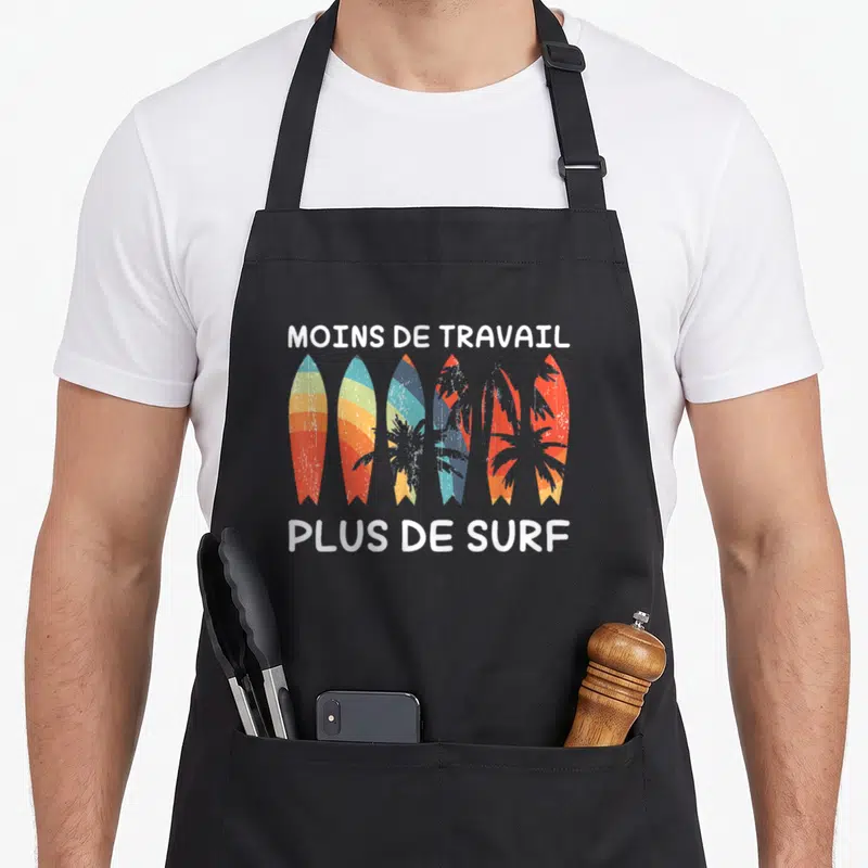 Tablier de cuisine noir avec six planches de surf colorées et le texte Moins de travail plus de surf en blanc.