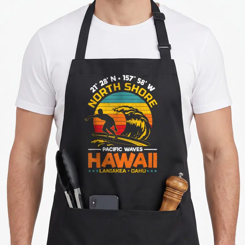 Tablier de cuisine noir avec illustration vintage d'un surfeur sur une vague orange devant un soleil couchant et le texte North Shore Hawaii.