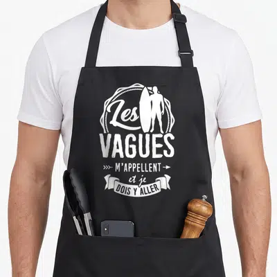 Tablier de cuisine noir avec silhouette de surfeur tenant une planche et texte Les Vagues m'appellent et je dois y aller en blanc.