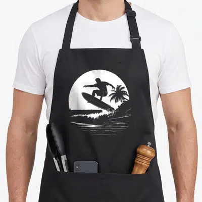 Tablier de cuisine noir avec silhouette blanche d'un surfeur sur une planche, devant un palmier et des vagues dans un cercle.